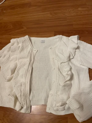 Vit kofta med volanger från Lindex - Superfin vit stickad kofta från Lindex med volangdetaljer längs framstycket och knappar hela vägen. Koftan har långärmade ribbstickade ärmar och rund hals. Perfekt för dig som gillar en söt och trendig look.