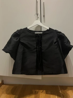 Svart blus med puffärm och snörning - Säljer en svart blus med korta puffärmar och snygg snörning framtill. Blusen har en rak och lite boxig passform, tillverkad i ett glansigt material som ger en cool look. Perfekt för dig som gillar statement-ärmar och trendiga detaljer.