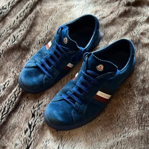 Blå mocka sneakers från Moncler - Snygga blå sneakers från Moncler i mocka med blå snörning och platt sula. Skorna har Moncler-logga på plös och häl, samt röd, vit och blå randig detalj på sidan. Perfekta för dig som gillar klassisk men sportig stil.