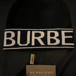 Burberry Mössa - Snygg svart mössa från Burberry med stor vit logotyp runt hela kanten. Mössan är stickad och har en klassisk, rund form som passar perfekt för kalla dagar. Materialet är mjukt och håller dig varm samtidigt som du får en stilren look.