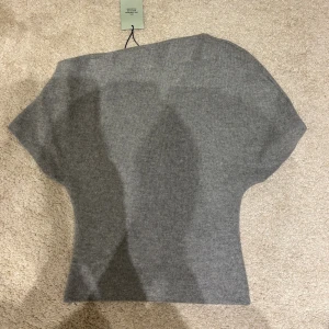 Grå stickad topp från Gina tricot  - Säljer en snygg grå stickad topp från Gina tricot med unik asymmetrisk halsringning och korta ärmar. Modellen är slim och sitter tajt över midjan, perfekt för dig som gillar stilrena och moderna plagg. Supermjuk och skön kvalitet! Storlek S, helt ny med prislapp! 