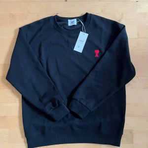 Svart sweatshirt från Ami Paris - Svart sweatshirt från Ami Paris med rund hals och långa ärmar. Tröjan har ett rött broderat hjärta med A på bröstet och ribbade muddar vid ärmslut och nederkant. Tillverkad i mjuk bomull för skön komfort. Perfekt för en chill och stilren look.