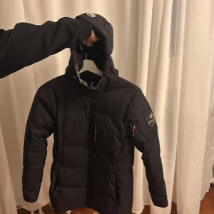 Svart pufferjacka från Sail Racing - Svart pufferjacka från Sail Racing med huva och dragkedja framtill. Jackan har logga på bröstet och ärmen, samt en orange detalj på ärmfickan. Långärmad och med quiltad design som ger en schysst look. Perfekt för kalla dagar och har en snygg, lite längre passform. 