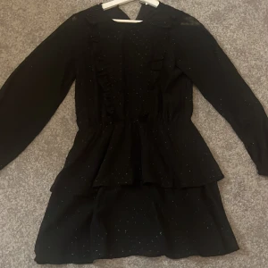 Svart glittrig volangklänning - Svart kortklänning med glittriga prickar och volangdetaljer från Gina Young. Klänningen har långa ärmar och är perfekt för en lite finare middag eller firande. Har bara använts ett fåtal gånger då jag hann växa ur den innan den var användbar. Säljer för 200 kr i storlek 158-164.💕