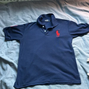 Mörkblå Polo kortärmad piké M - Snygg mörkblå pikétröja från Polo i 100% bomull. Klassisk krage med två knappar och rött broderat Polo-logga på bröstet. Kortärmad modell med röd detalj på ärmen. Perfekt för en chill och stilren look.