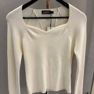 Vit ribbad tröja från Soaked - Supersnygg vit ribbad tröja från Soaked med lång ärm och unik fyrkantig halsringning. Stickad struktur med diskreta mönsterdetaljer framtill ger en modern vibe. Perfekt att styla med jeans eller kjol för en clean look.