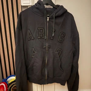 Axel Arigato hoodie - Svart hoodie med dragkedja från Axel Arigato. Tröjan har stora broderade bokstäver framtill, huva med snörning och en klassisk känguruficka. Långärmad och perfekt för chill dagar. Snygg och stilren design med mörka detaljer. Herr modell