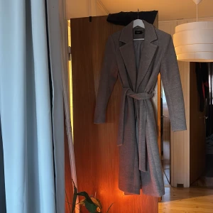 Grå/beige kappa med bälte från ONLY - Snygg grå/begie kappa från ONLY i storlek S. Modellen har klassisk krage, långa ärmar och ett matchande bälte i midjan för en clean siluett. Insidan är fodrad i ljusgrått tyg och kappan har en rak, lite längre passform som ger en stilren vibe.