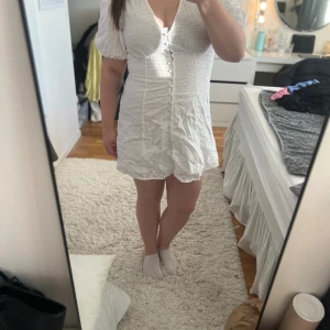 Vit kort klänning från Gina Tricot - Supersöt vit kort klänning från Gina Tricot med broderade blommor, puffärmar och v-ringning. Klänningen har knappar framtill och är gjord i bomull. Perfekt för sommarens alla tillfällen, och studenten. Ena bandet till en av knapparna har gått sönder men det är inget man tänker på, men utöver det så är den helt ny och prislappen är fortfarande kvar!! Den är i storlek 42 men skulle mer säga att det är en 38-40!!💕✨