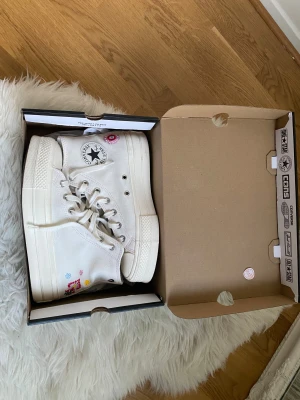 Converse vita höga sneakers med broderi - Använda 3-4 gånger. Säljer ett par vita Converse All Star sneakers med högt skaft och coola broderade blommor i flera färger på sidan. Skorna har snörning, rund tå och platt sula. Materialet är klassisk canvas och de har den ikoniska Converse-loggan på skaftet.