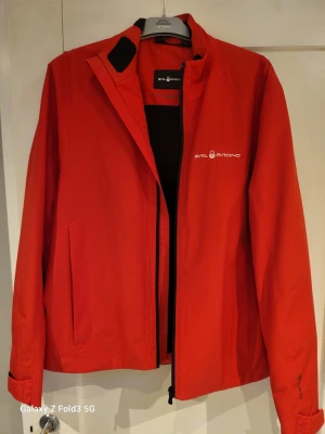 Röd Gore-Tex vindjacka Sail Racing - Snygg röd vindjacka från Sail Racing i storlek medium. Jackan är tillverkad i Gore-Tex-material som håller dig torr och har svart fleecefoder på insidan. Den har dragkedja framtill, diskret logga på bröstet och justerbara ärmslut med kardborre. Perfekt för blåsiga dagar.