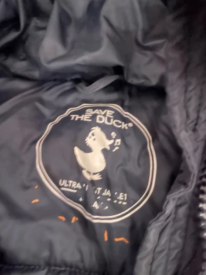 Mörkblå  lång pufferjacka Save The Duck - Säljer en mörkblå, lång pufferjacka från Save The Duck med huva och snygg quiltad design. Jackan har dragkedja framtill och ett orange logomärke på ärmen. Perfekt för kalla dagar och har en clean, modern look.