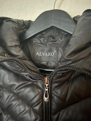 Dunjacka Alvaro - Svart pufferjacka från Alvaro med snyggt leopardmönstrat foder. Jackan har huva, dragkedja framtill och två fickor med dragkedja. Materialet är glansigt och lätt, perfekt för kyliga dagar. Jackan har en figurnära passform och långa ärmar. Använd en del men ingen synlig slitage som finns på jackan. Väldigt varm och fiin.
