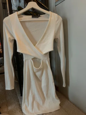 Vit omlottklänning från PrettyLittleThing - Supersnygg vit omlottklänning från PrettyLittleThing med lång ärm och ribbad struktur. Klänningen har en djup v-ringning och cool cut-out detalj vid midjan som ger en trendig vibe. Perfekt för dig som gillar stilrena och moderna plagg.