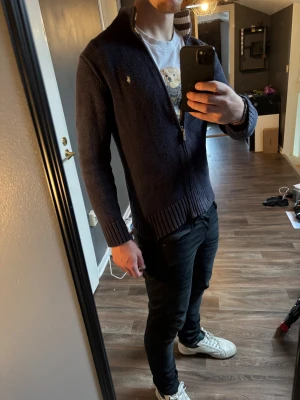 Marinblå kofta från Ralph Lauren - Säljer en marinblå stickad kofta från Ralph Lauren med dragkedja och ribbade muddar. Koftan har en ståkrage med randig insida och den klassiska broderade loggan på bröstet. Perfekt för lager-på-lager och snygg till jeans. Det är i barnstorlek men sitter som S.