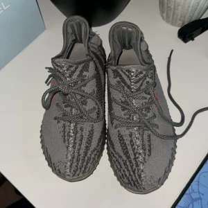 Adidas Yeezy Boost 350 V2 Beluga - Säljer ett par Adidas Yeezy Boost 350 V2 Beluga med grå och mörkgrå ränder, orange 'SPLY-350' text på sidan och ribbad platt sula. Skorna har snörning och är tillverkade i stickad syntet med en mjuk boost-sula för extra komfort. Storlek 37