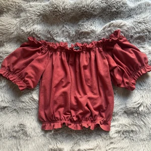 Röd offshoulder topp - Säljer en röd offshoulder topp från Gina Tricot med korta puffärmar och volangdetaljer vid halsringning och nederkant. Croppad passform som sitter snyggt över axlarna. Perfekt för dig som gillar trendiga och feminina plagg.