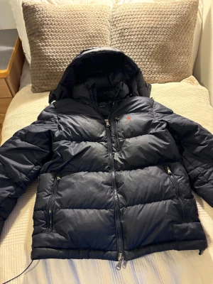 Svart dunjacka från Polo Ralph Lauren - Snygg svart dunjacka från Polo Ralph Lauren med huva och röd broderad logga på bröstet. Jackan har två sidofickor med dragkedja och en kraftig tvåvägsdragkedja framtill. Perfekt för kalla dagar och har en puffad look som är riktigt trendig. Jackan är en xs men passar mig som har M i kläder
