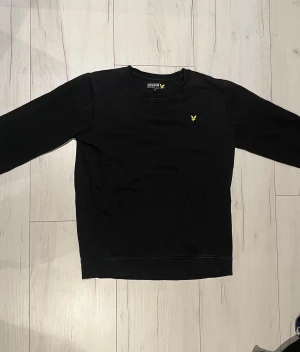 Svart sweatshirt från Lyle & Scott - Svart sweatshirt från Lyle & Scott med klassisk rund hals och gul broderad logga på bröstet. Tröjan har långärmad passform och är gjord i mjuk bomull, perfekt för chill dagar. Enkel och stilren design som funkar till jeans eller joggers.