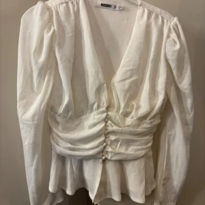 Vit blus  - Säljer en vit blus från Missguided med puffiga långärmar och djup v-ringning. Blusen har knappar framtill och snygg rynkning i midjan som ger en cool siluett. Materialet är strukturerat och känns lätt mot huden. Perfekt för dig som gillar statement-ärmar och trendiga detaljer.