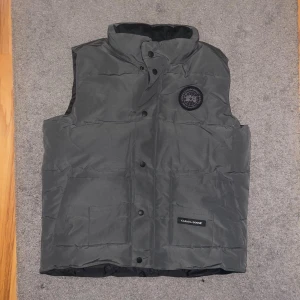 Mörkgrå väst från Canada Goose - Snygg mörkgrå väst från Canada Goose med hög krage och svart logotyp-patch på bröstet. Västen har tryckknappar framtill, två stora fickor och diskret Canada Goose-märke på ena fickan. Perfekt för kalla dagar och har en clean, urban look.