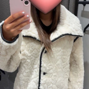 Vit teddyjacka från Gina Tricot - Säljer en fluffig vit teddyjacka eftersom jag köpt den i fel storlek, från Gina Tricot med stora svarta knappar och kontrastdetaljer längs sömmarna. Jackan har bred krage, lång ärm och loose passform. Perfekt för dig som vill ha en mysig och trendig look i vinter. 🥰