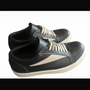 Svarta sneakers från Rick Owens - Snygga svarta sneakers från Rick Owens med vit kontrastdetalj på sidan och vita snören. Skorna har en rund tå, platt vit sula och är tillverkade i skinn med inslag av mocka. Kommer med originalkartong och extra svarta skosnören.