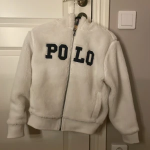 Vit teddyjacka från Polo Ralph Lauren - Säljer en fluffig vit teddyjacka från Polo Ralph Lauren med stor POLO-text i mörkblått på bröstet. Jackan har dragkedja framtill, ribbade muddar och mjukt foder. Äkta. Nyskick. Endast använd ett fåtal gånger. Perfekt julklapp.
