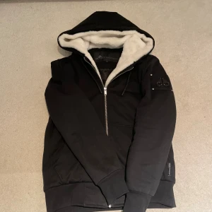 Svart Moose Knuckles jacka - Svart hoodiejacka från Moose Knuckles med vit päls i huvan och quiltat innerfoder. Jackan har dragkedja framtill, ribbade muddar och en snygg logga på ärmen. Perfekt för kalla dagar med sin varma och mjuka insida.