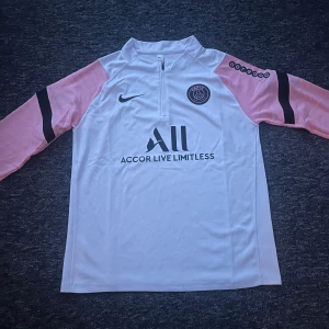PSG långärmad fotbollströja Nike - Paris Saint-Germain långärmad träningströja från Nike i vitt med ljusrosa ärmar och svarta detaljer. Tröjan har halv dragkedja, PSG-logga på bröstet och sponsortryck fram och bak. Tillverkad i lätt och ventilerande funktionsmaterial.
