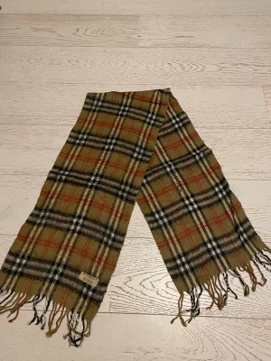 Rutig halsduk från Burberry i ull - Klassisk rutig halsduk från Burberry i 100% lammull. Beige bas med svarta, vita och röda rutor, samt fransar i ändarna. Perfekt accessoar för att lyfta din outfit med ikonisk brittisk stil.