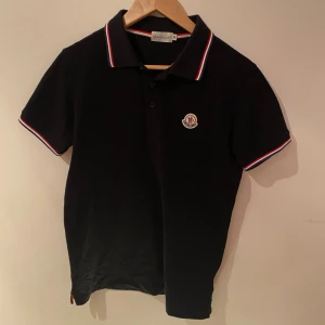 Svart Moncler pikétröja med röd/vit kant - Svart pikétröja från Moncler med korta ärmar och klassisk krage. Tröjan har Moncler-logga på bröstet och snygga rödvita detaljer på krage och ärmslut. Perfekt för dig som gillar stilrena och sportiga plagg med exklusiv känsla.