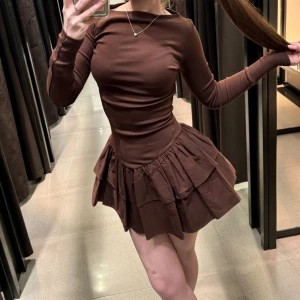 Brun volangklänning med lång ärm - Supersnygg brun kort klänning med långärmad överdel och volangkjol i flera lager. Klänningen har en tight passform upptill och en fluffig kjol som ger en cool siluett. Perfekt för dig som gillar att sticka ut med en unik look. Materialet känns mjukt och stretchigt.