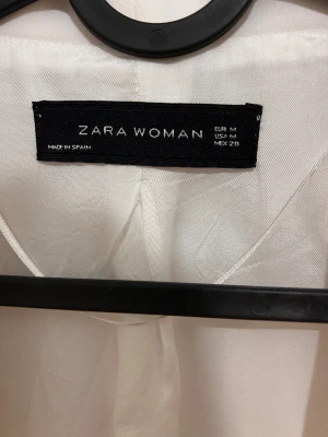 Vit kavaj från Zara Woman, M - Snygg vit kavaj från Zara Woman i storlek M. Stilren design med klassisk krage, två fickor framtill och lätt uppvikta ärmar. Materialet är mjukt och har en lätt glansig finish som ger en fräsch look. Perfekt för att lyfta din outfit.
