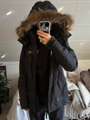 Svart parkajacka från Abercrombie L - Parkajacka från Abercrombie & Fitch i svart med fluffig fuskpäls runt huvan och broderad A&F-logga på bröstet. Jackan har dragkedja, knappar och flera fickor framtill. Insidan är fodrad med mjuk teddy och materialet är slitstark polyester.