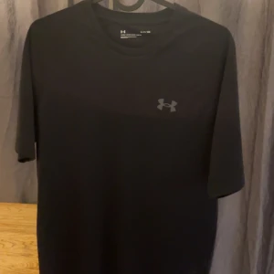 Svart Under Armour t-shirt i polyester - Säljer en svart t-shirt från Under Armour med diskret logga på bröstet. T-shirten har korta ärmar och är gjord i mjukt, lätt polyester som andas. Perfekt för dig som gillar sportig och enkel stil. Passformen är normal och materialet känns skönt mot huden.