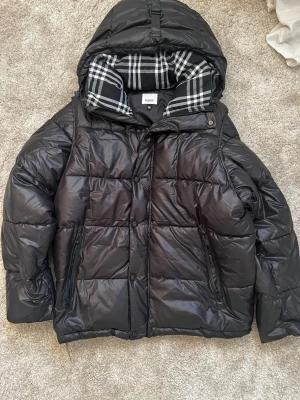 Svart pufferjacka från Burberry - Säljer en svart pufferjacka från Burberry med huva och klassiskt rutigt foder. Jackan har en patch med Burberry London England på ärmen, dragkedja och knappar framtill samt två sidofickor med dragkedja. Perfekt för kalla dagar. Den är även en convertivle till en väst. Säljer den eftersom den är för stor för mig.