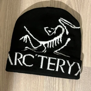 Svart Arcteryx mössa med logga - Snygg svart mössa från Arcteryx med stor vit logga och text framtill. Perfekt för dig som gillar streetwear och vill ha något som sticker ut. Materialet är stickat och mössan har uppvikt kant för extra stilpoäng.
