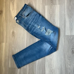 Blå jeans från Jack & Jones - Snygga blå skinny jeans från Jack & Jones, modell Liam. Jeansen har slitna detaljer och hål på ena benen för en cool look. Klassisk femficksdesign och smal passform som sitter skönt längs benen. Perfekta för dig som gillar mode.