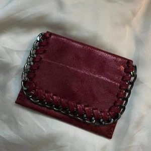 Vinröd plånbok med kedjedetalj - Snygg vinröd plånbok med glansig finish och cool kedjedetalj runt kanten. Plånboken har flera fack för kort och stängs med tryckknapp. Perfekt storlek för att ha med sig överallt och ger en edgy vibe till din stil.