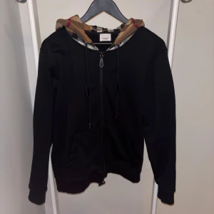 Svart Burberry hoodie med rutig huva - Svart hoodie från Burberry med dragkedja och klassisk rutig huva i beige, svart, vit och röd. Tröjan har snörning vid huvan, två fickor framtill och ribbade muddar. Perfekt för dig som vill ha en stilren look med en touch av lyx.