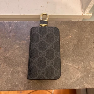 Svart GG plånbok  - Snygg svart plånbok från GG med klassiskt GG-mönster och guldfärgade detaljer. Plånboken har dragkedja runtom och en praktisk nyckelring inuti. Perfekt att hänga på väskan eller använda som nyckelfodral.