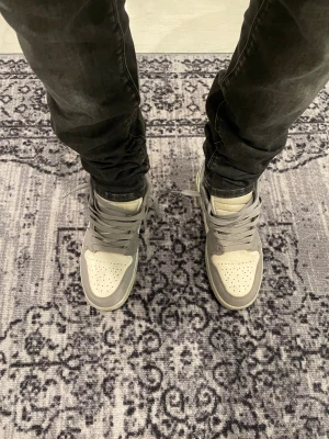 Nike Air Jordan 1 Low grå/vit - Nike Air Jordan 1 Low sneakers i grått och vitt med klassisk Jumpman-logga på tungan. Skorna har snörning, rund tå och platt sula. Materialet är en mix av skinn och syntet, med perforeringar på tån för extra ventilation. Snygg och stilren färgkombination som passar till streetwear.