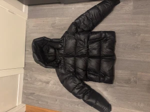 Svart pufferjacka från J.Lindeberg - Säljer en svart pufferjacka från J.Lindeberg med huva och dragkedja framtill. Jackan har en quiltad design och är tillverkad i syntetmaterial med dunfyllning. Märkespatch på ärmen och två sidofickor med dragkedja. Perfekt för kalla dagar. Storlek S men passar m bättre 