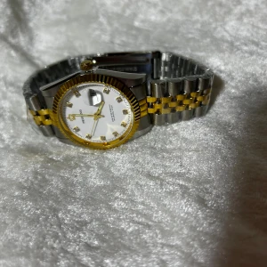 Proking silver och gul klocka med Datum  - Snygg klocka från Proking med rund vit urtavla, guldfärgade detaljer och markerade index med stenar. Armbandet är i silver och guld, med klassisk länkdesign i metall. Klockan har datumvisning och guldfärgad krona. Perfekt för dig som gillar stilrena accessoarer.