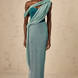 Turkos omlott långklänning med slits - Klänning från miss rosier. Snygg turkos långklänning med omlott-detalj och knyt över axeln. Klänningen har en glittrig yta och oneshoulder-design som ger en cool och unik look. Materialet är lätt och skimrande, med en slits bak för extra rörelsefrihet. Perfekt för dig som vill sticka ut. Helt ny oanvänd. 