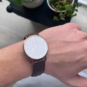 Daniel Wellington klocka med brun läderrem - Snygg och stilren klocka från Daniel Wellington med rund vit urtavla, guldiga detaljer och klassisk brun läderrem. Vattentät 3 ATM. Behöver bytas batteri
