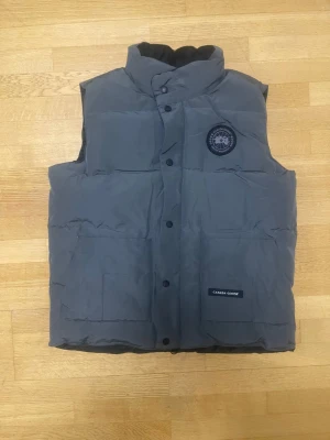 Canada Goose väst - Snygg grå dunväst från Canada Goose med hög krage och svart logotyp på bröstet. Västen har stora fickor framtill, knappar och diskret Canada Goose-märke. Perfekt för kalla dagar när du vill hålla stilen och värmen.