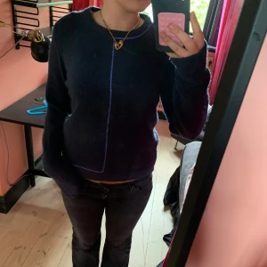 Mörkblå stickad tröja med lila kant - Mysig mörkblå stickad tröja med lång ärm och rund hals. Tröjan har en unik lila linje som går längs framsidan och runt halsen, vilket ger en cool och modern look. Perfekt för dig som gillar stilrena men ändå speciela plagg.