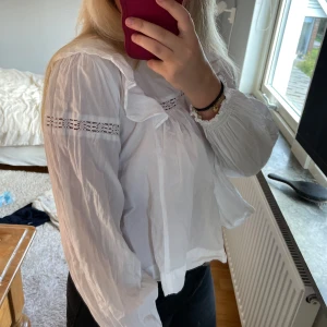 Vit blus med volang från Gina Tricot - Supersöt vit blus från Gina Tricot med volangdetaljer och spetsinslag vid axlar och bröst. Blusen har långärmade puffärmar med resår vid ärmslut och en lätt v-ringning. Tillverkad i bomull och har en luftig, loose passform.